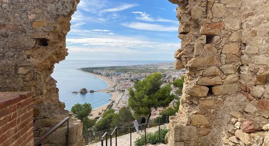 Vista desde el **castillo de Blanes** hacia la bahía y la ciudad. El mar azul contrasta con las ruinas de piedra y el cielo nublado.