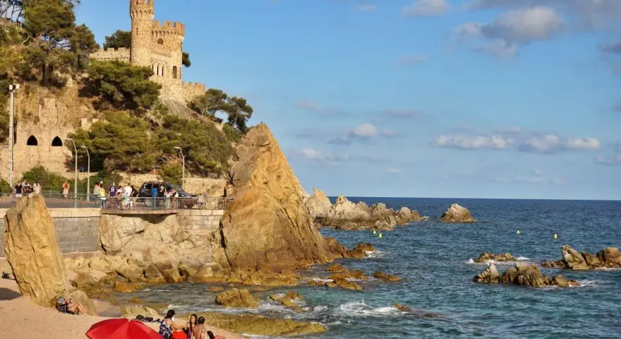 Castillo medieval sobre acantilados rocosos en sa caleta Lloret de Mar. Playa con gente y mar azul.