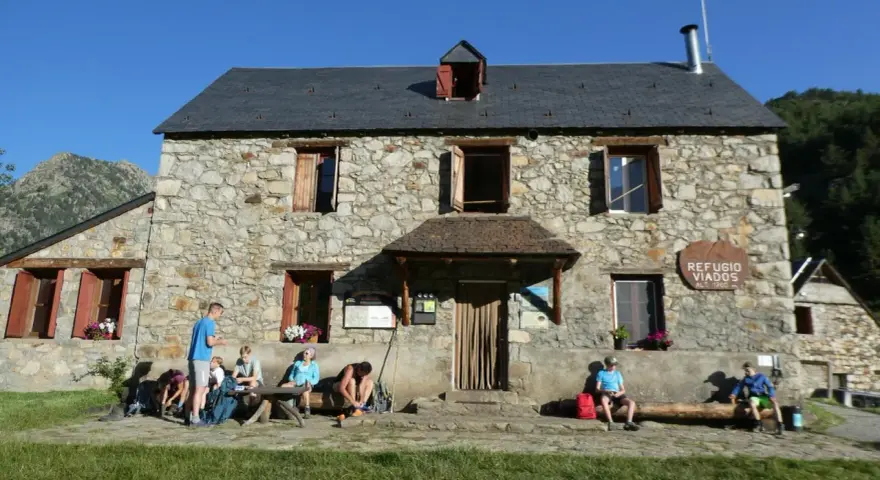 Grupo de excursionistas descansa frente al Refugio Viados, parte de la ruta de los tres refugios. Montañas y cielo azul al fondo.