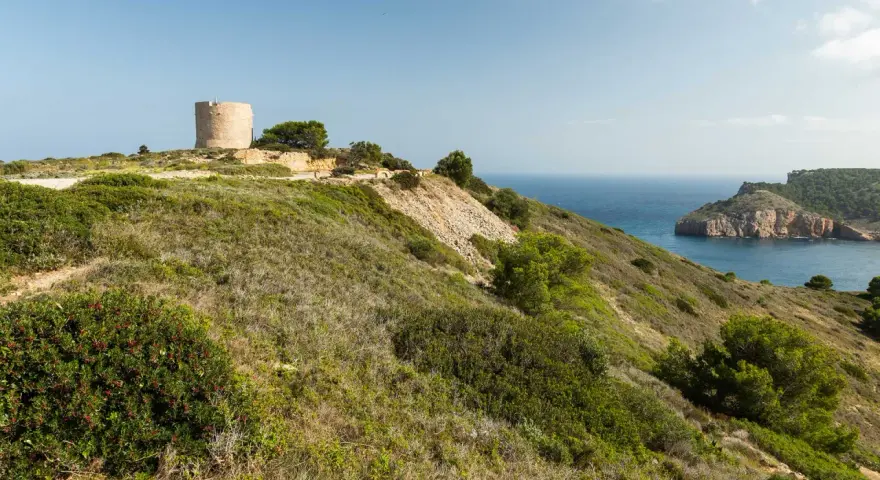 La torre de Montgó se alza sobre una colina verde, vigilando el mar azul y una bahía rocosa.