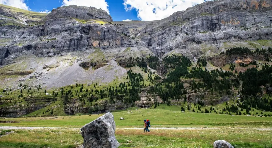 Senderista camina por un prado verde con imponentes montañas de **3000 del Pirineo** al fondo.