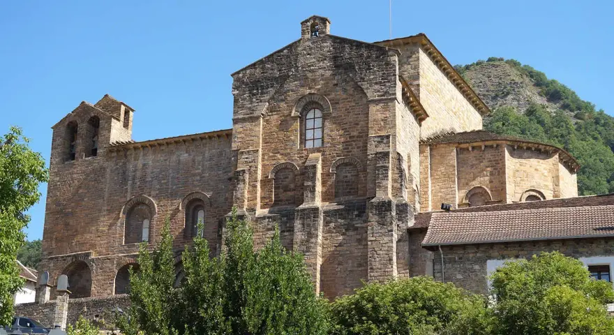 El **monasterio de Siresa**, con su imponente arquitectura de piedra y campanario, se alza bajo un cielo azul, rodeado de vegetación exuberante.