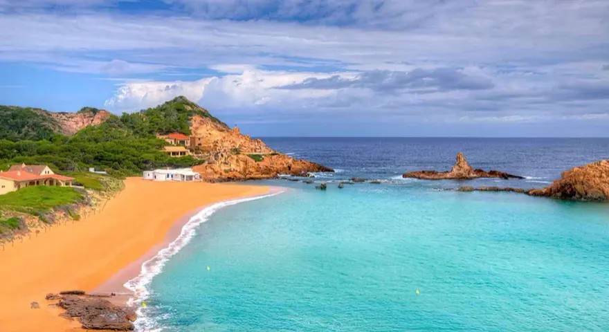 Hermosas calas cerca de Fornells, con arena dorada, aguas turquesas y casas blancas integradas en la costa rocosa.