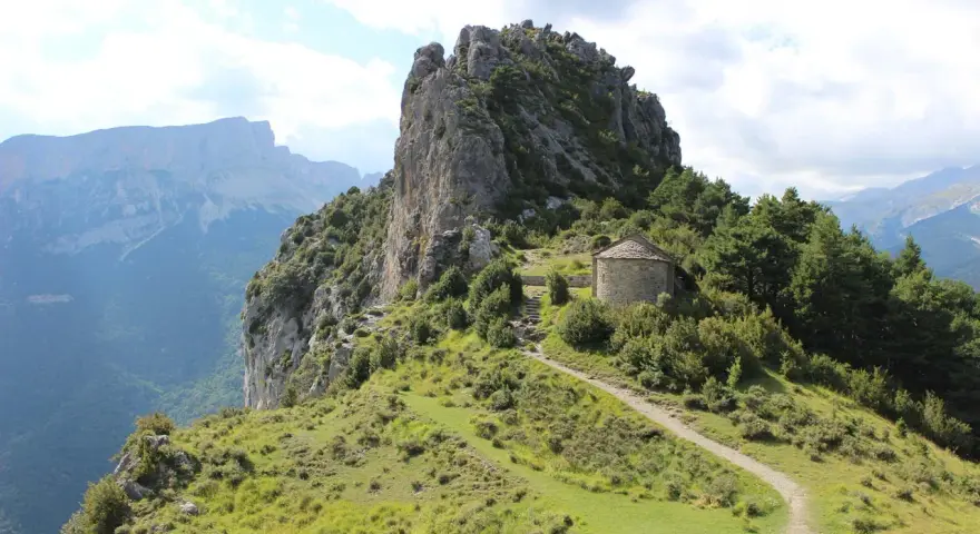 Pequeña ermita de piedra con tejado cónico, rodeada de vegetación y un sendero que sube por la ladera. Las ermitas de Tella se alzan majestuosas.