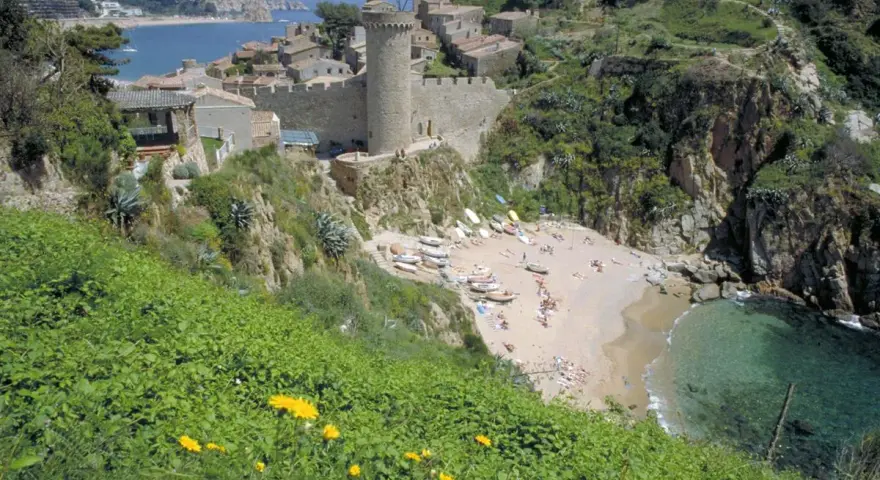 Vista de las calas de Tossa de Mar, con un castillo medieval y una playa con gente disfrutando del sol y el mar.