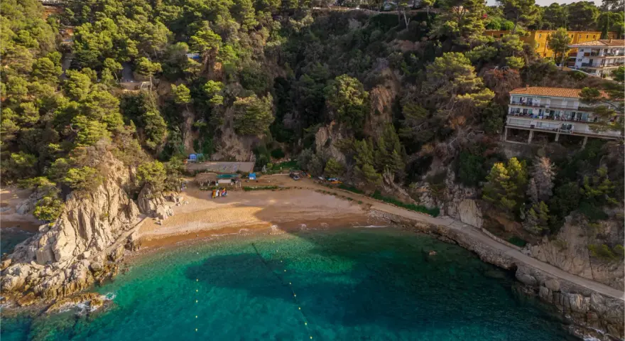Bahía de Cala Bona, Barcelona, con aguas turquesas, arena dorada y acantilados verdes. Casas en la ladera.