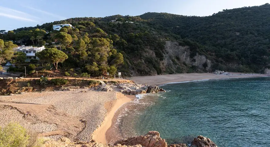 La platja de Canyerets, con su arena dorada y aguas cristalinas, se extiende bajo una colina verde y casas blancas.