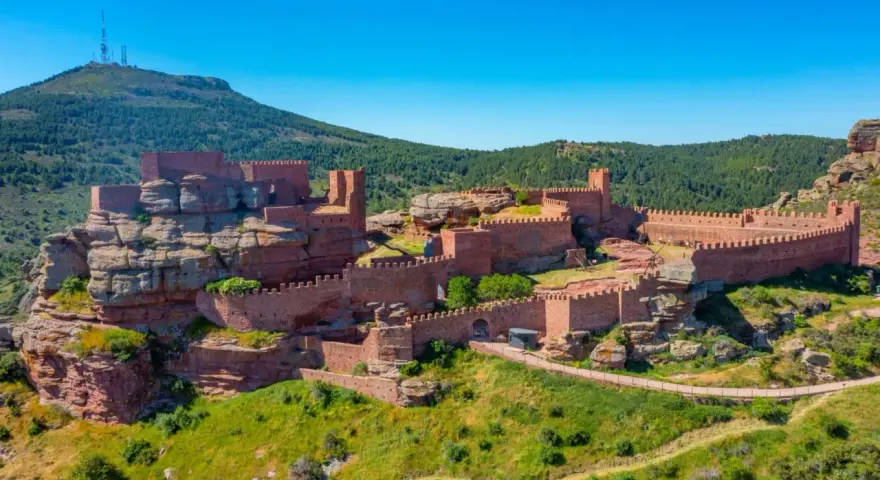 Imponente castillo de Peracense, una fortaleza de piedra rojiza que se alza sobre un peñón, rodeada de vegetación y bajo un cielo azul intenso.