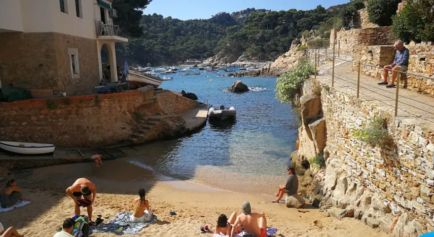 Gente relajándose en la arena de cala Malaret, Begur, con barcos anclados en aguas cristalinas y colinas verdes al fondo.