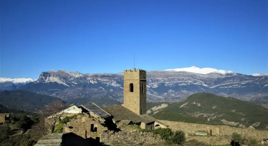 Antiguo muro de roda con torreón de piedra y ruinas, rodeado de montañas nevadas bajo un cielo azul intenso.