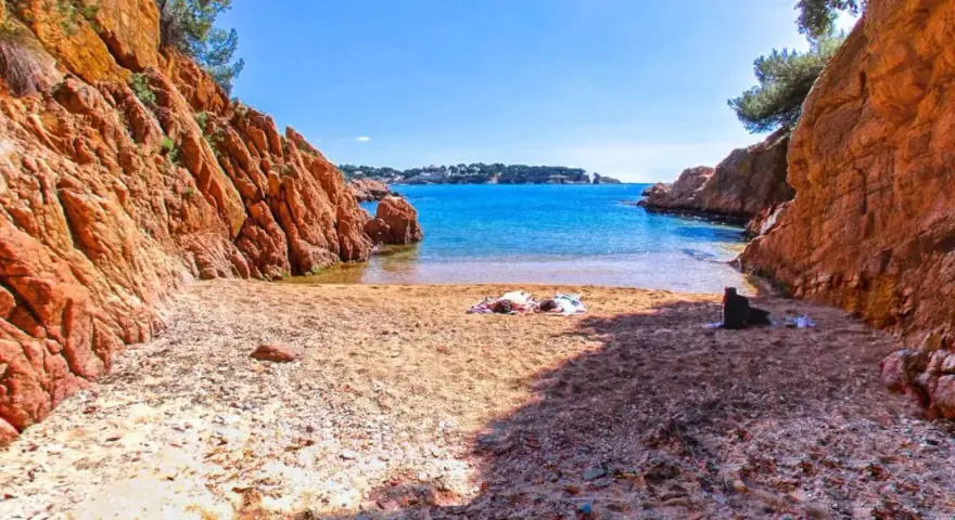 Arena dorada y aguas turquesas en la cala Maset. Dos personas toman el sol tranquilamente en la playa, rodeadas de imponentes rocas rojizas.