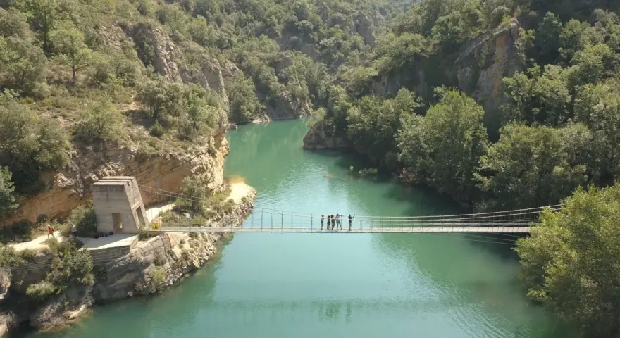 Un puente colgante cruza un río turquesa rodeado de montañas verdes. Un grupo de personas camina por el puente de Montañana.