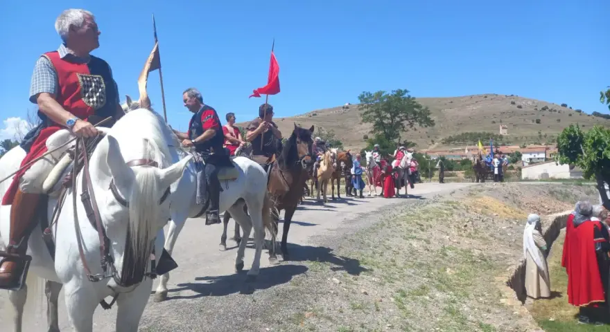 Un desfile ecuestre con trajes históricos, quizás recreando la gesta de el poyo del cid, avanza por un camino rural bajo un cielo azul.