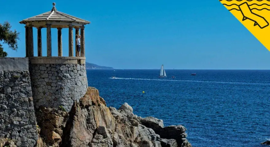Un mirador de piedra con columnas se alza sobre rocas, con el mar azul y veleros al fondo. Un lugar perfecto para pasear por los **caminos de ronda**.
