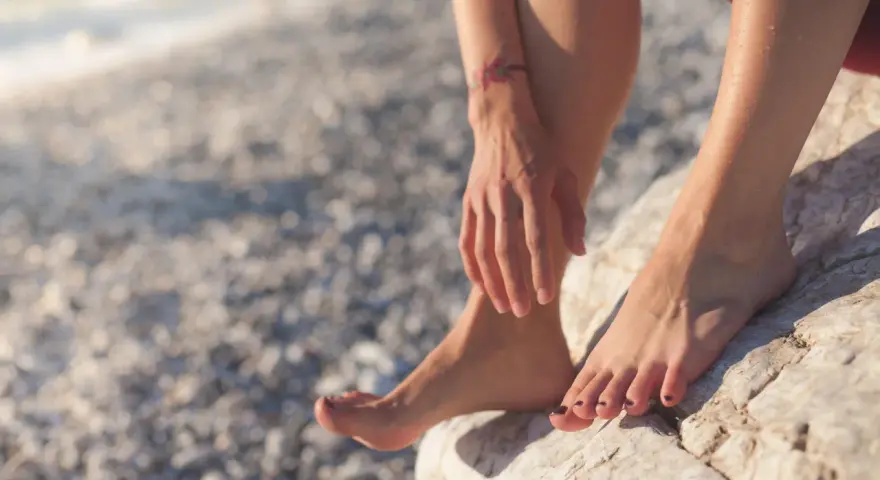 Pies descalzos sobre rocas en una playa nudista de Cadaqués. El sol brilla sobre la piel mojada y un pequeño tatuaje de flores adorna el brazo.