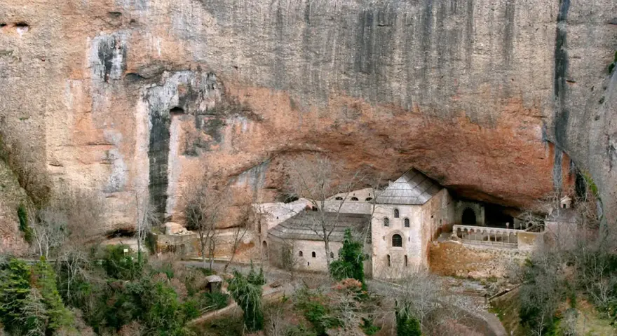 Monasterio de San Juan de la Peña, un impresionante conjunto arquitectónico excavado en la roca, con edificios de piedra y tejados a dos aguas bajo un gran saliente rocoso.