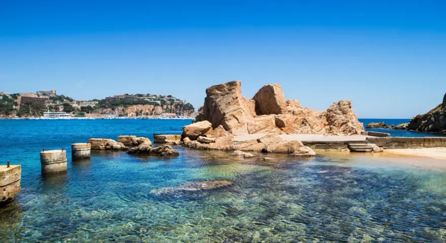 Un paseo marítimo en Sant Feliu de Guíxols, con rocas doradas y aguas cristalinas. El pueblo se alza en la colina.