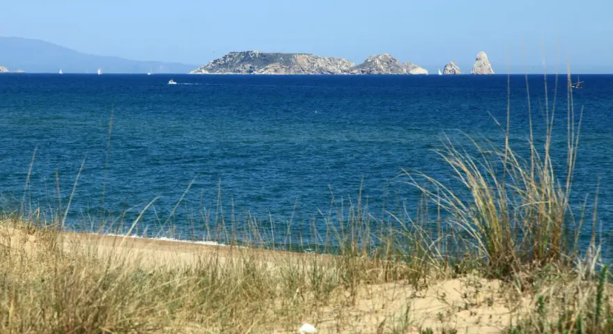 Dunas de hierba seca en primer plano, mar azul y un archipiélago rocoso en la distancia, evocando la belleza de la playa mas Pinell.