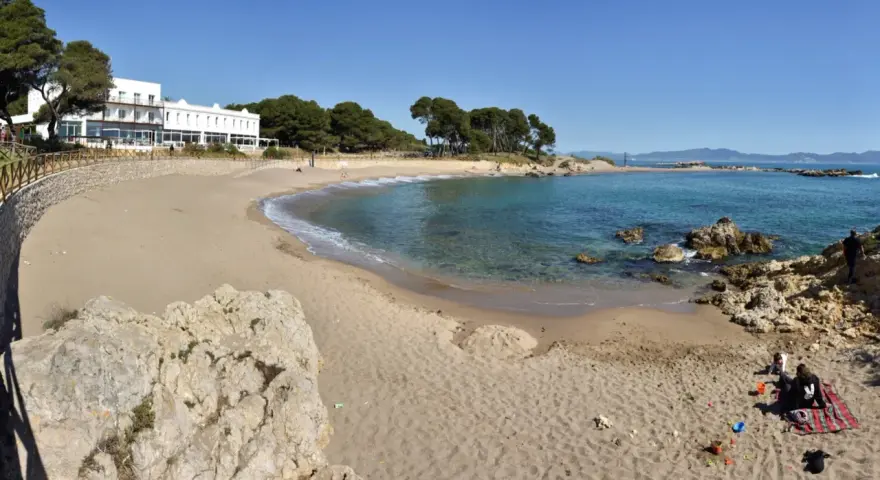 Platja Cala de la Creu: arena dorada, aguas turquesas y un hotel blanco con vistas al mar. Familias disfrutan del sol y juegan en la arena.