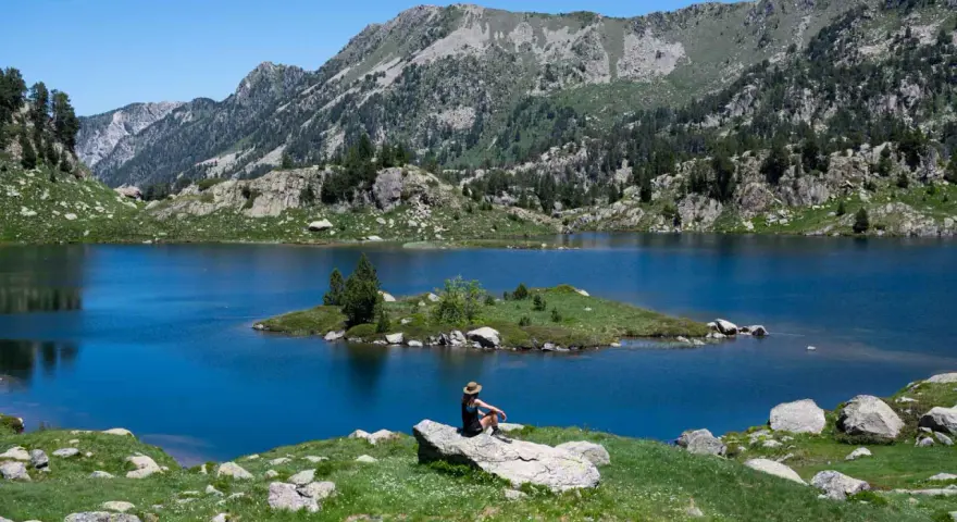 Persona sentada en roca contempla los lagos de Colomers en una ruta corta. Paisaje montañoso y azul intenso.