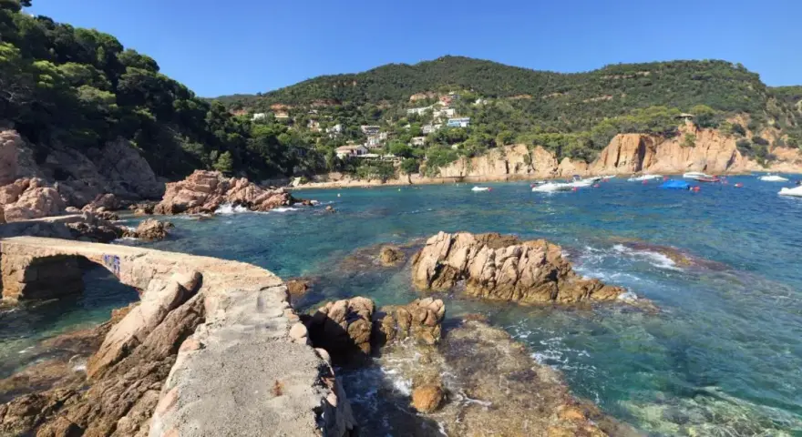 Un puente de piedra cruza aguas turquesas en la cala Canyet. Barcas flotan tranquilas mientras colinas verdes y casas blancas coronan el paisaje.