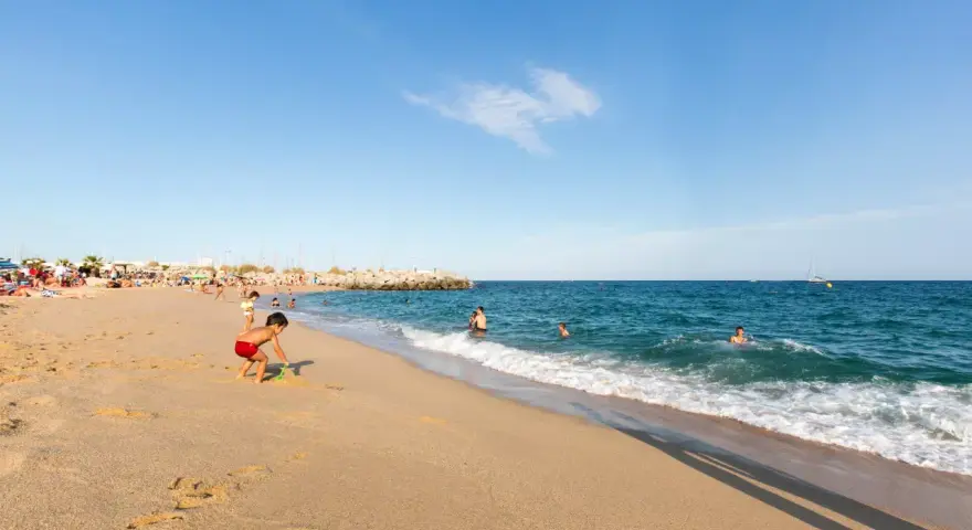 Un niño juega en la arena dorada de la playa de les barques, mientras otros disfrutan del mar azul y un velero navega en el horizonte.