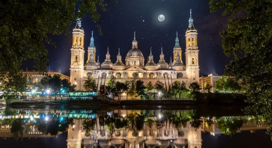 La majestuosa basílica del Pilar, iluminada bajo un cielo estrellado y la luna, se refleja en el río.