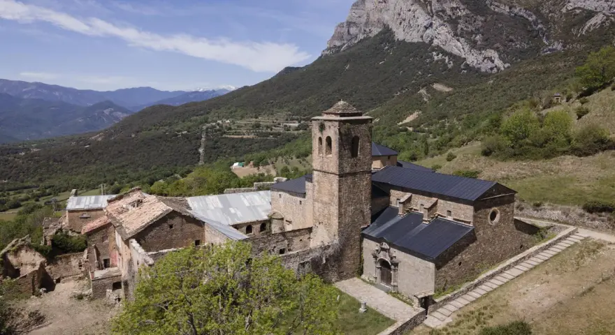 El monasterio de San Victorián, un conjunto de edificios de piedra con tejados oscuros, se alza majestuoso ante una imponente montaña rocosa.