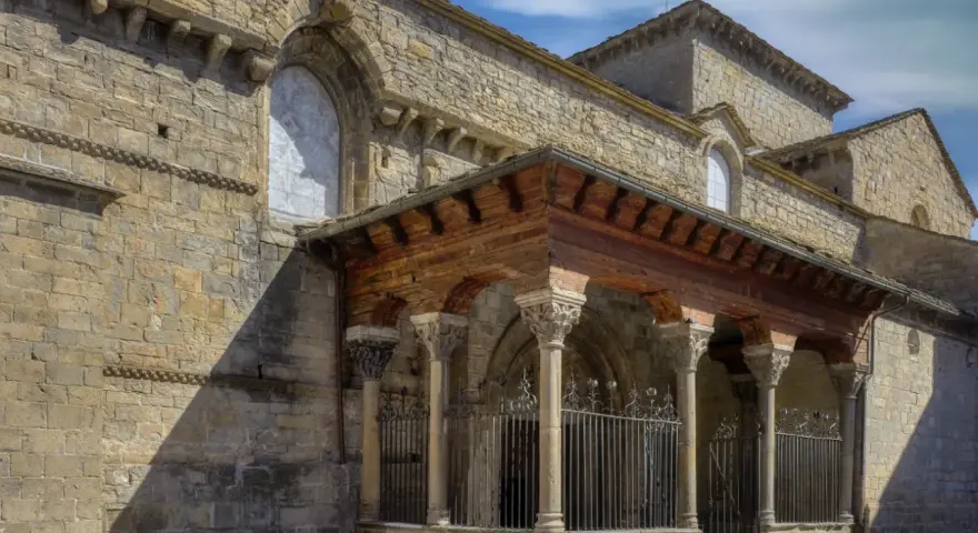 La **catedral de Jaca**, con su imponente arquitectura románica, se alza bajo un cielo azul. Un pórtico de madera y piedra protege la entrada.