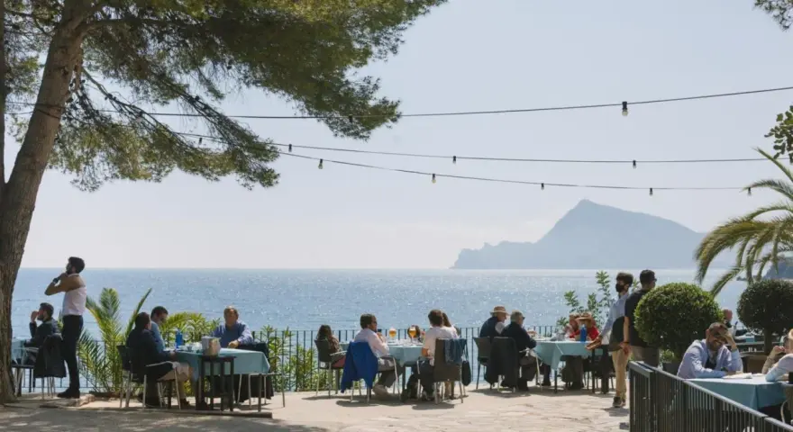 Gente disfrutando de una comida al aire libre con vistas al mar. Un lugar ideal donde comer en Altea, con mesas azules y un ambiente relajado bajo un árbol.
