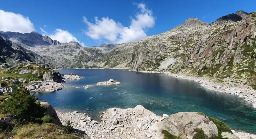 Lago de aguas cristalinas rodeado de montañas rocosas en el Vall de Boí. El cielo azul con nubes blancas contrasta con el verde de la vegetación.