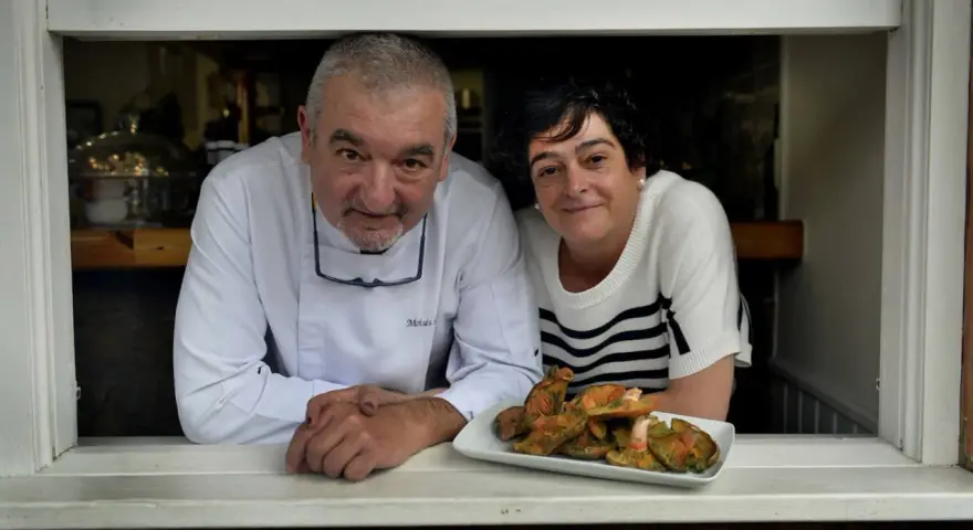 Chef y camarera sonríen tras ventana con plato de setas. Descubre dónde comer en Castro Urdiales.