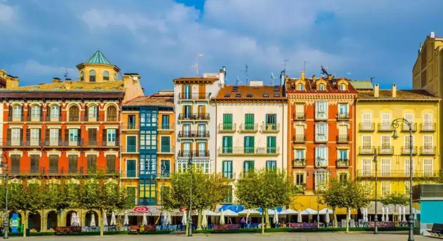 Fachada de edificios coloridos en una plaza, ideal para saber **qué ver en Pamplona**. Terrazas y árboles verdes bajo un cielo azul.