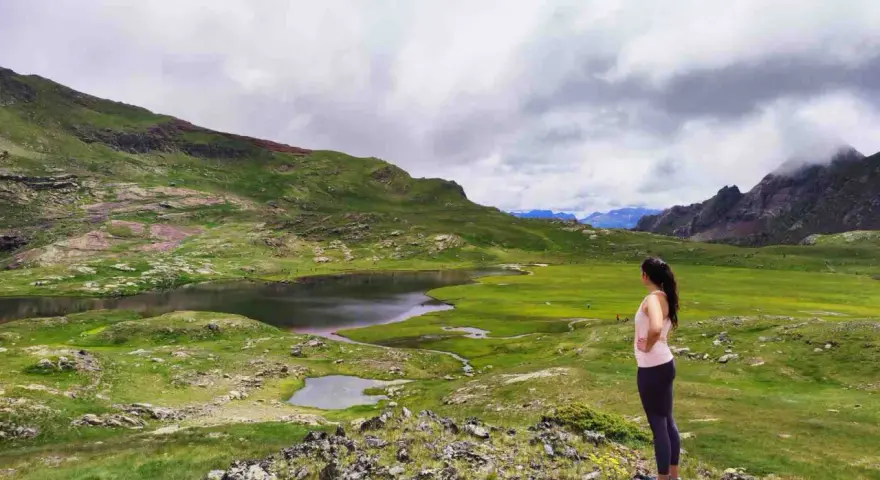 Mujer contempla los ibones del Valle de Tena, un paisaje de lagos y prados verdes bajo un cielo nublado.