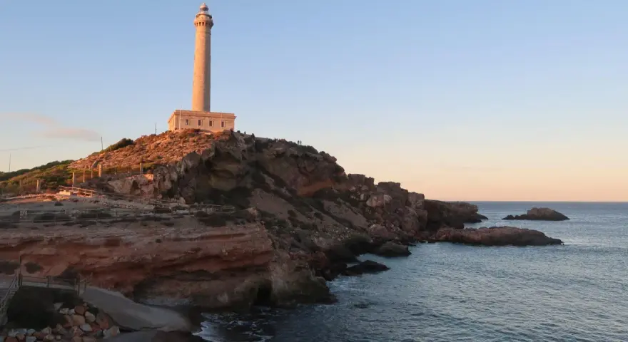 Faro sobre acantilados rojizos, con el mar azul y un cielo despejado. Un camino desciende hacia una pequeña cala roja.