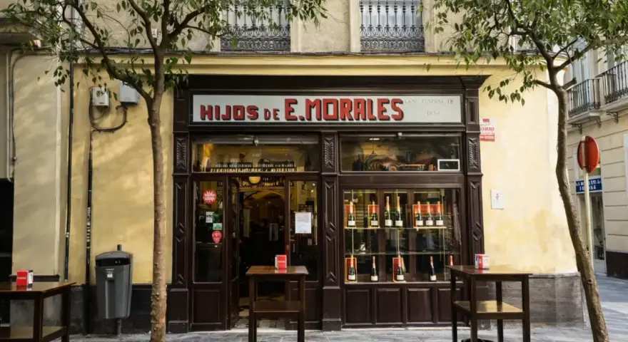 Fachada de "Hijos de E. Morales", un bar tradicional en Sevilla, con mesas en la acera y botellas expuestas.
