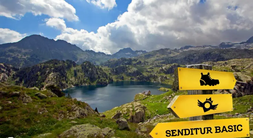 Paisaje de montaña con un lago azul, señalización de sendero y el letrero "SENDITUR BASIC", parte de la ruta de los 7 lagos Pirineos.