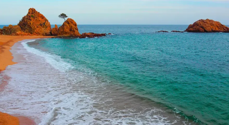 La platja mar menuda, con sus rocas anaranjadas y aguas turquesas, invita a soñar. Una ola rompe suavemente en la arena dorada.