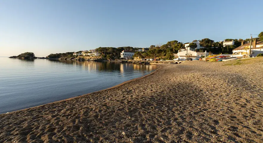 La platja gran de Cadaqués, con casas blancas y kayaks coloridos, se baña en la calma del mar al atardecer.