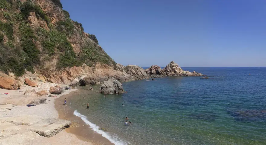 Les platges de Blanes ofereixen aigües cristal·lines i roques imponents sota un cel blau. Gent gaudeix del sol i del mar.