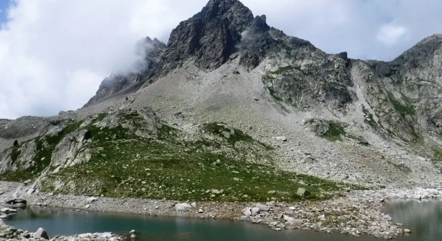 Paisaje montañoso con un lago de aguas tranquilas en primer plano. Las laderas están cubiertas de rocas y vegetación verde, con picos escarpados y nubes en el cielo. Los **ibones de Arriel** reflejan la majestuosidad del entorno.