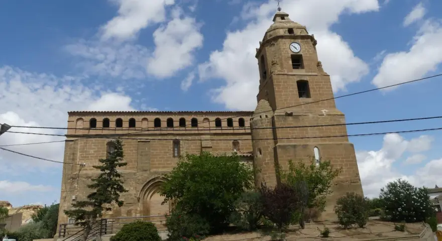 Iglesia de piedra con torre y reloj, rodeada de vegetación. Un día soleado en Alberuela de Tubo.