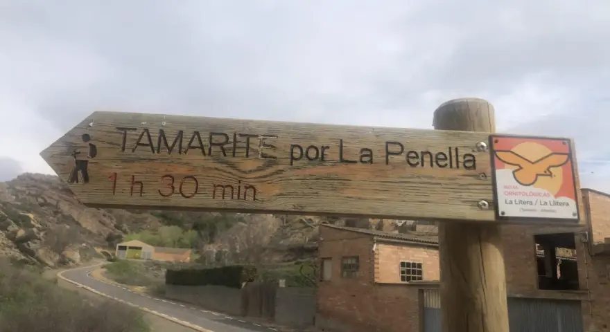 Señal de madera en albelda huesca: "Tamarite por La Penella, 1h 30 min". Al lado, señal de rutas ornitológicas.