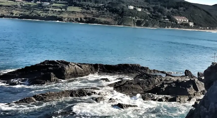 Olas rompiendo en rocas oscuras, con la costa verde y edificios de Mundaka al fondo. Un lugar perfecto para comer y disfrutar del paisaje.