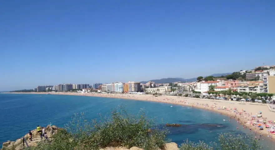 Vista panorámica de la playa S'Abanell en Blanes, con el mar azul intenso, arena dorada repleta de gente y edificios costeros bajo un cielo despejado.
