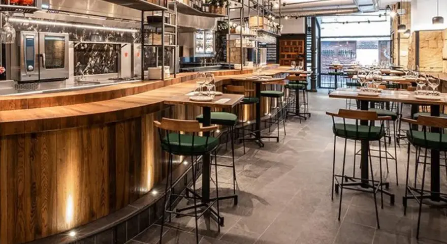 Bar moderno con taburetes verdes y mesas de madera. Un lugar ideal donde comer en Salamanca, con cocina a la vista y ambiente acogedor.