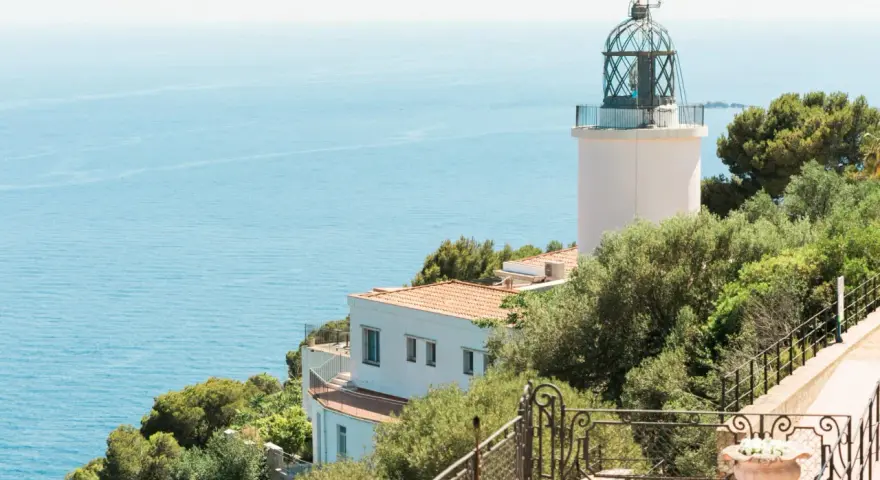 El faro de Sant Sebastià se alza sobre el mar azul, rodeado de vegetación exuberante.