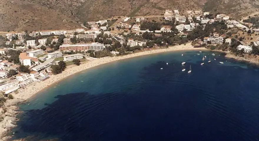 Vista aérea de la playa Almadrava en Roses, con edificios blancos y barcos anclados en aguas azules.