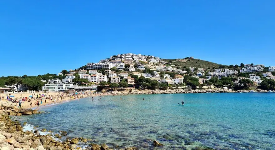 Vista de la platja de Montgó, con gente disfrutando del sol y el mar turquesa, y casas blancas escalonadas en la colina.
