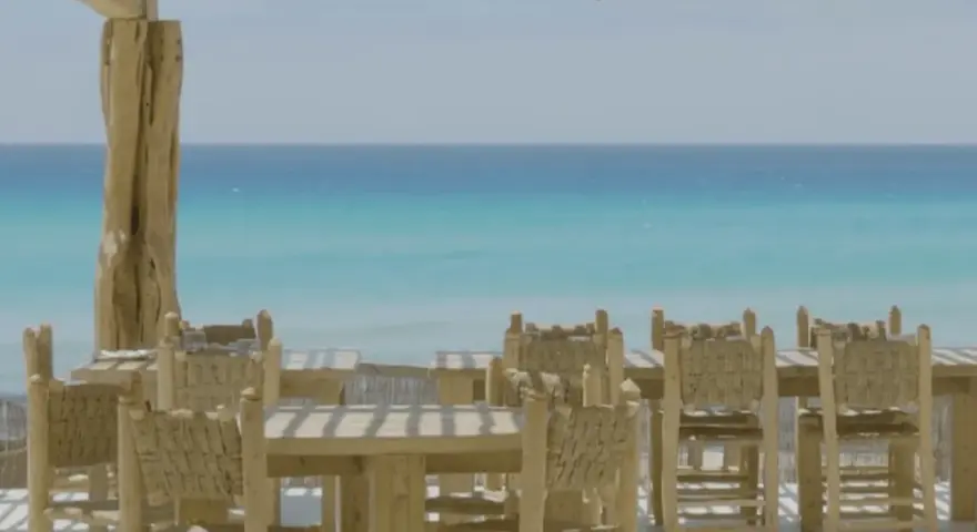 Mesas y sillas de madera rústica en la terraza de restaurantes en Formentera, con el mar turquesa de fondo.